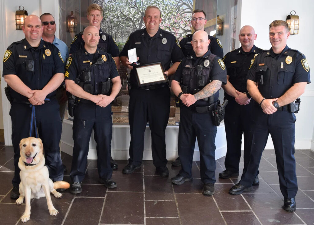 A group of police officers, including a dog (Officer Zane) stand in the lobby of Town Hall. The man in the middle holds a citation and a medal.
