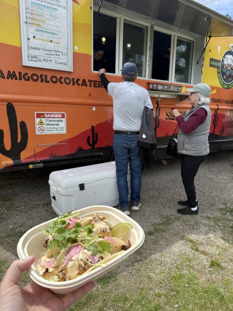 In the foreground, a paper bowl with tacos toped with pickled onions and cilantro. In the background, tow people order from truck that has cacti painted on the side.