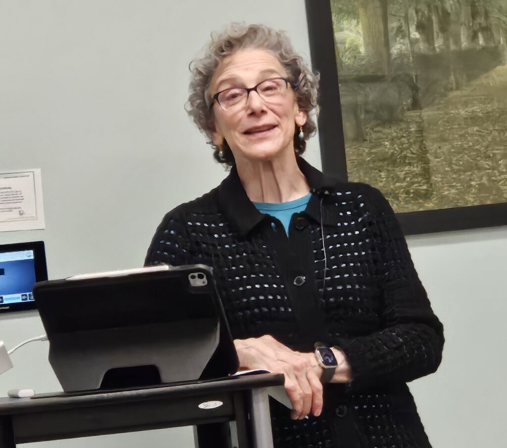 A curly-haired woman in a black sweater leans against a podium.