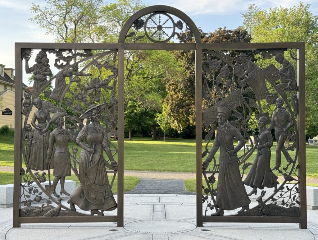 A gate in the middle of a park. Pictures of women from different historical eras decorate the gate.