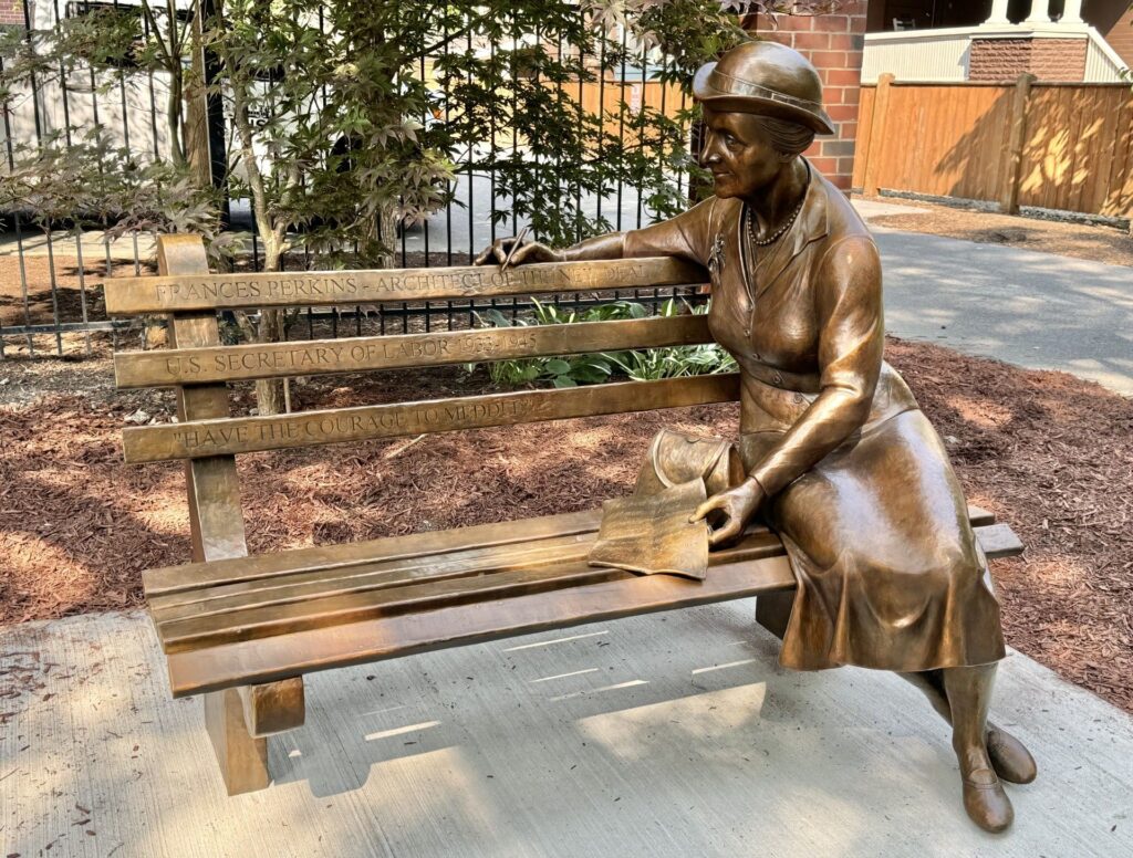 A full-sized sculpture of a woman sitting on a park bench. The bench has words carved into it and the woman holds a pice of paper and wears an old-fashioned hat.