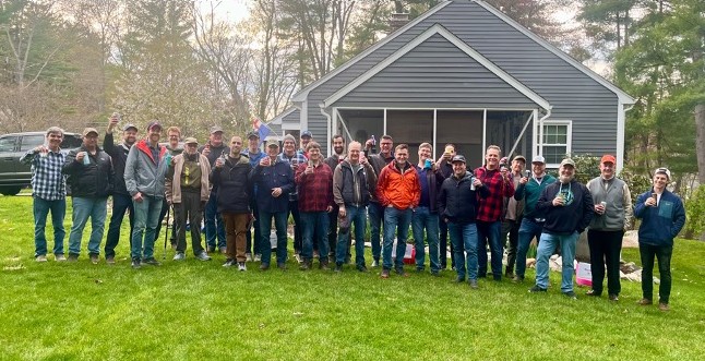 A large group of men in front of a house. Each person is holding a can of beer.