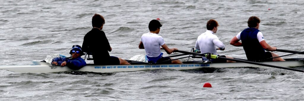 Four rowers push hard while the coxswain watches from the boat well.