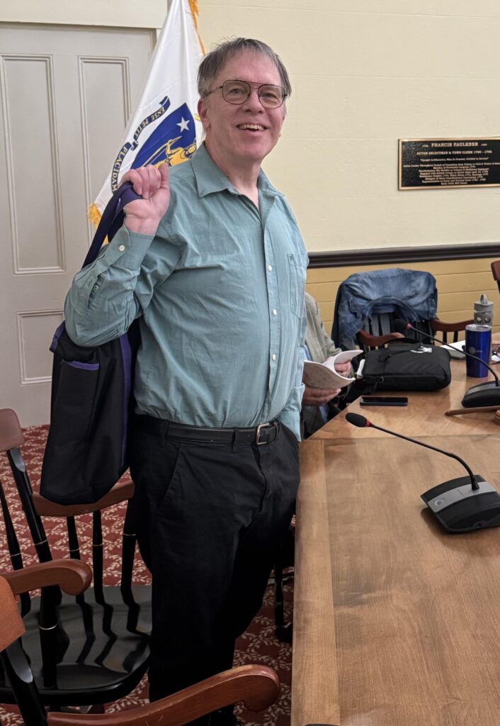 A man wearing alarge grin and a wrinkly shirt (as usual) stands at a desk in Room 204.