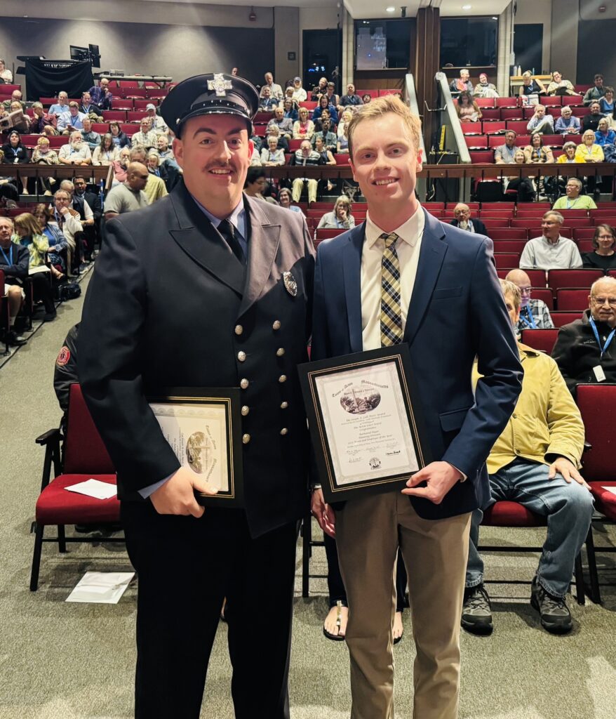 Two men stand in front of an audience holding citations. The man on the left is wearing a firefighter dress uniform (with two rows of brass buttons!) and the young man on the right wears a jacket and plaid tie.