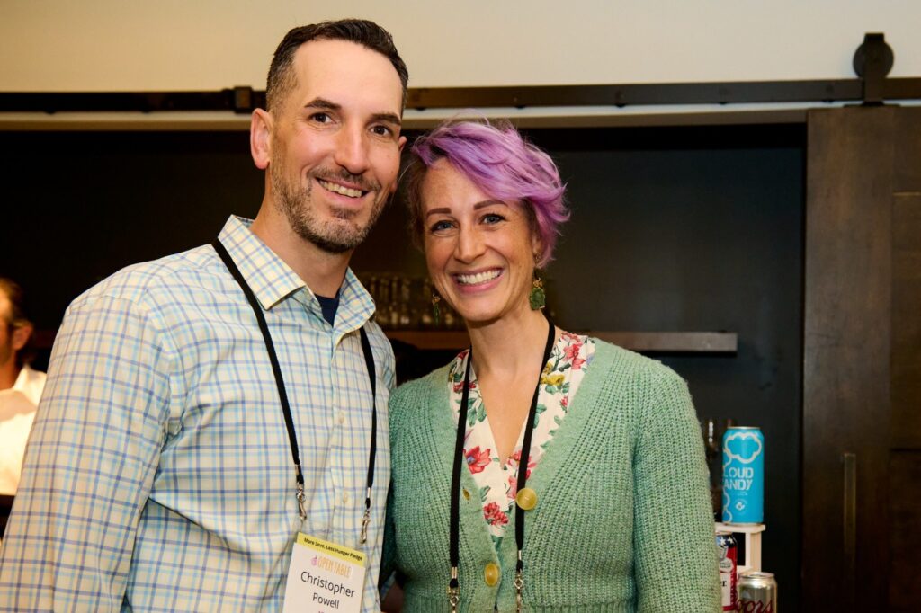 Two smiling people stand in front of a bar. The woman is sporting lilac-colored hair and a green sweater.