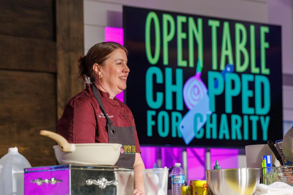A chef stands in front of a sign that says "Open Table Chopped for Charity," and behind a work table with a frypan, metal bowls, and other cooking implements.