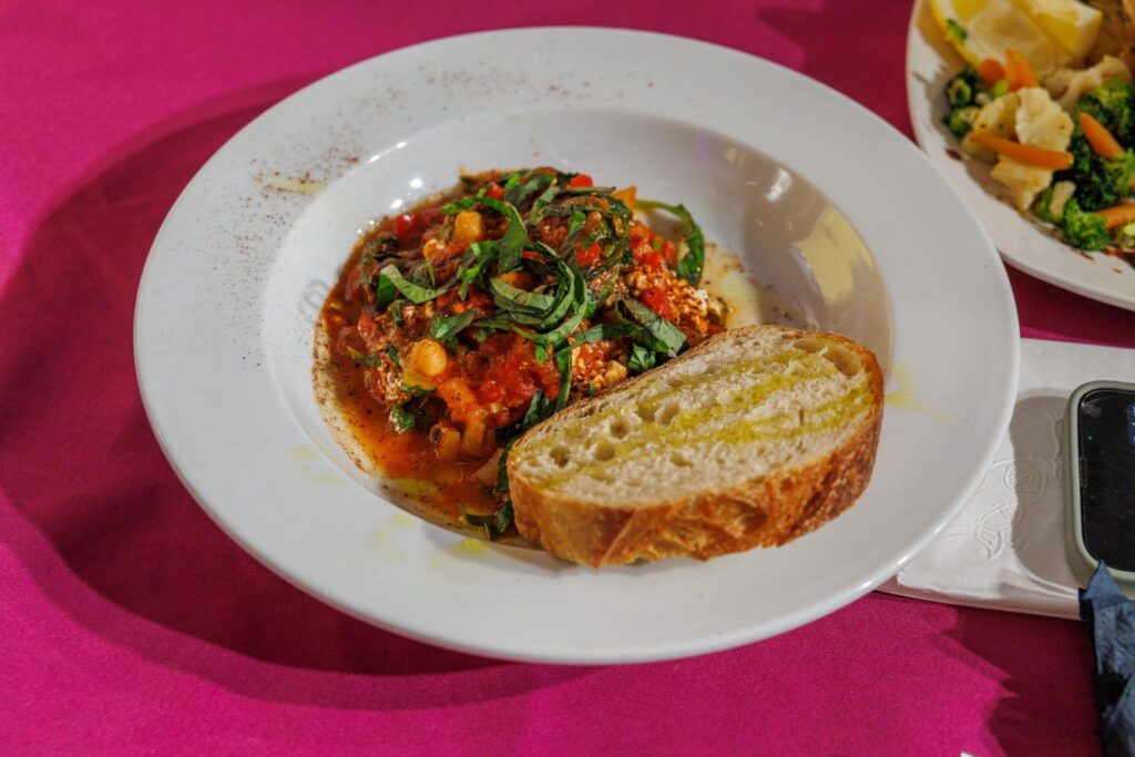 A white shallow boal with a dollop of a tomato and chickpea stew is accompanied by a slice of olive-oil drizzled bread.