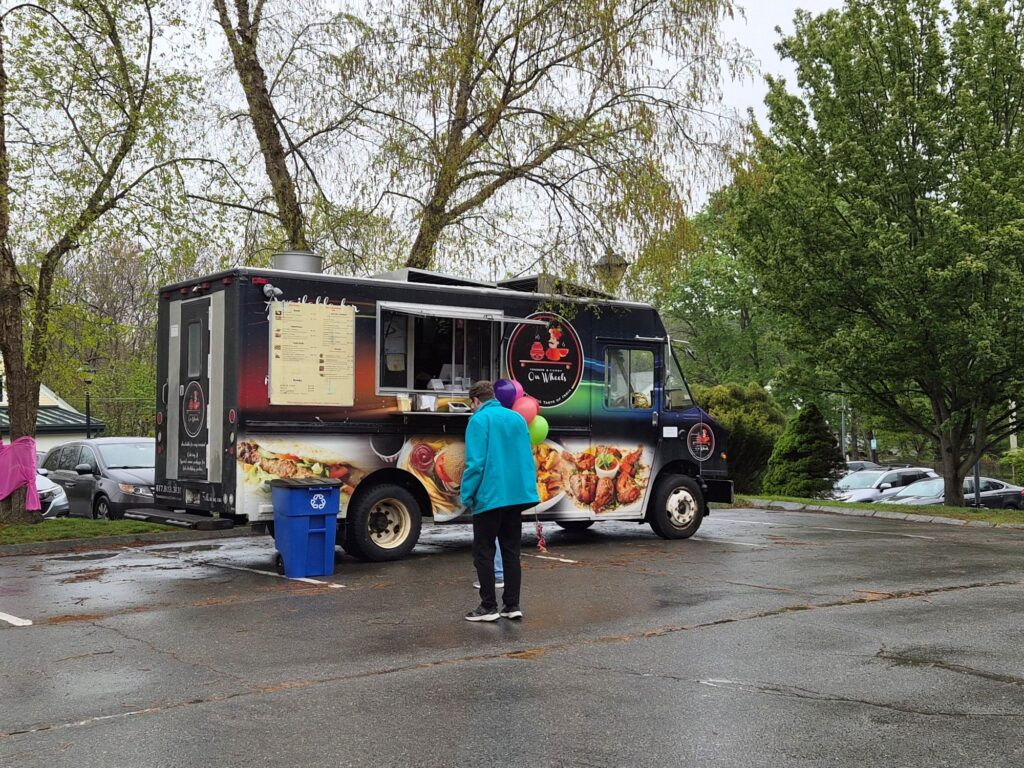 A food truck stands in a damp parking lot. A few people are in line.