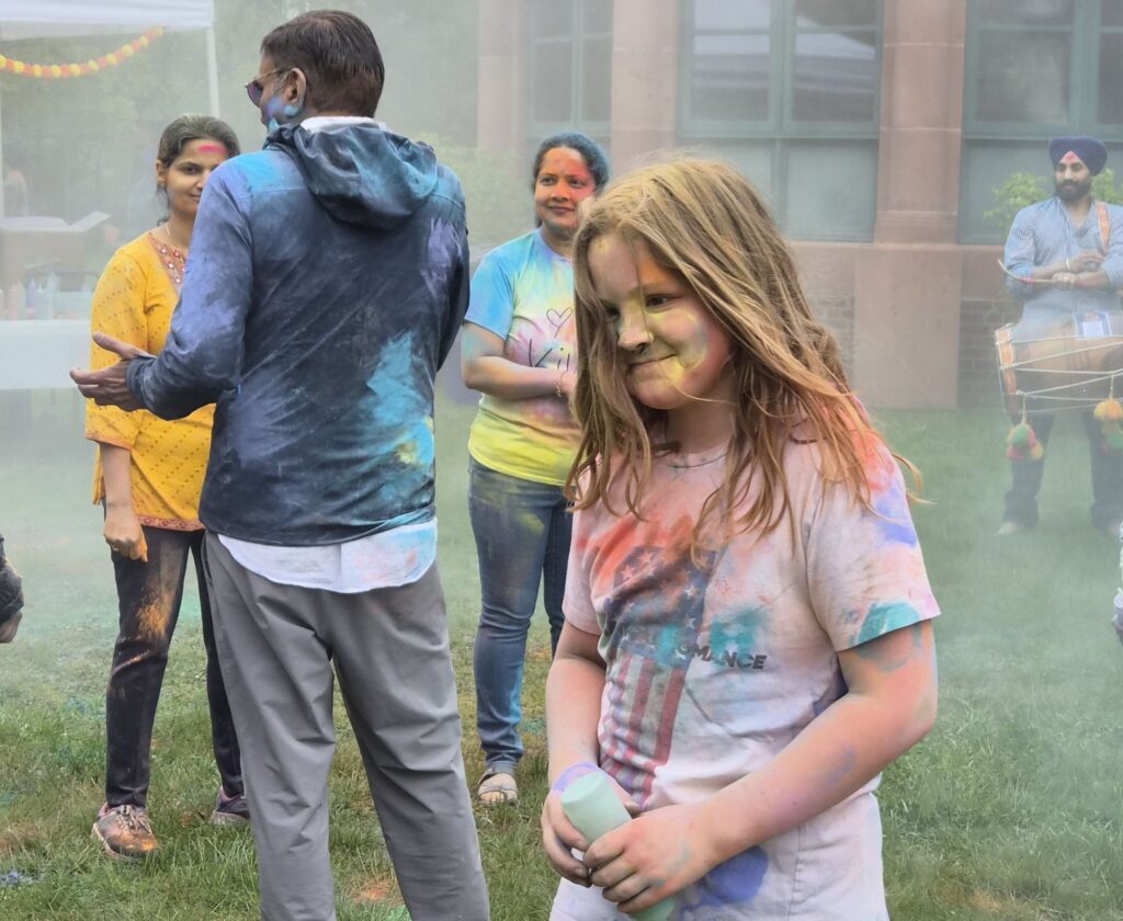 A child in need of a shower. A kid with shoulder length hair and a white t-shirt, all of which are splotched with many colors. He holds a squeeze bottle in his hands and seems to be heading towards someone. In the background, a few adults are chatting -- also covered in color.
