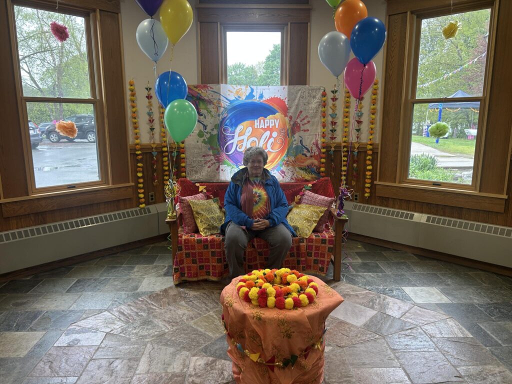 A quiet spot in the lbrary. In front of the window is a bench decorated with red and gold fabric and pillows, balloons, and other decorations. A decorated table is in the foreground. An older woman in a tie-dye t-shirt sits on the bench.