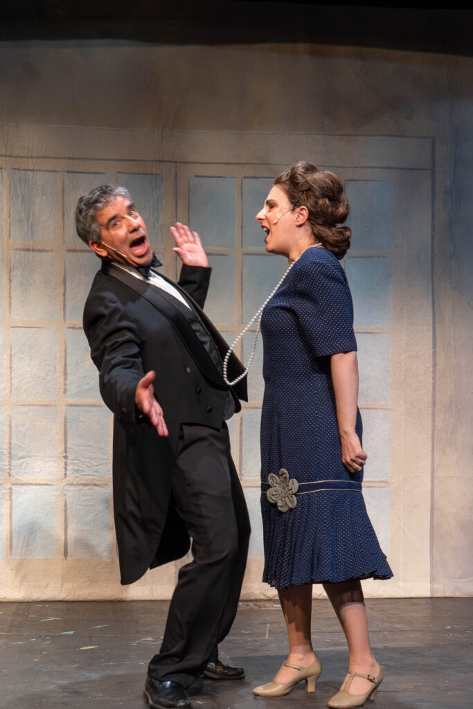 A man in formal tails leans away from a woman in a blue dress (with a swinging pearl necklace). They are both singing.