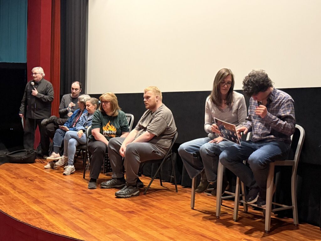 Four pairs of people sit on the stage in front of the screen. One young man is using an alphabet card to say something to his aide.
