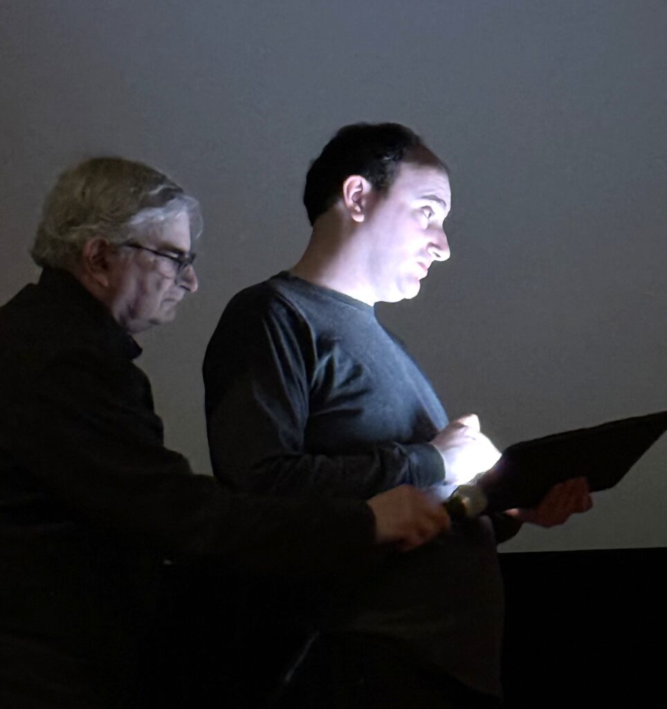 An older man on the laft stands behind a taller young man. The older man is looking at an iPad (or something like it).