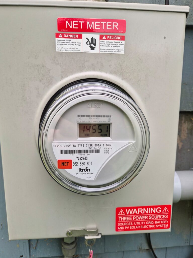 A home outdoor power meter. A large grey box iwth a window that shows some numbers. There are a handful of red warning stickers on the meter.