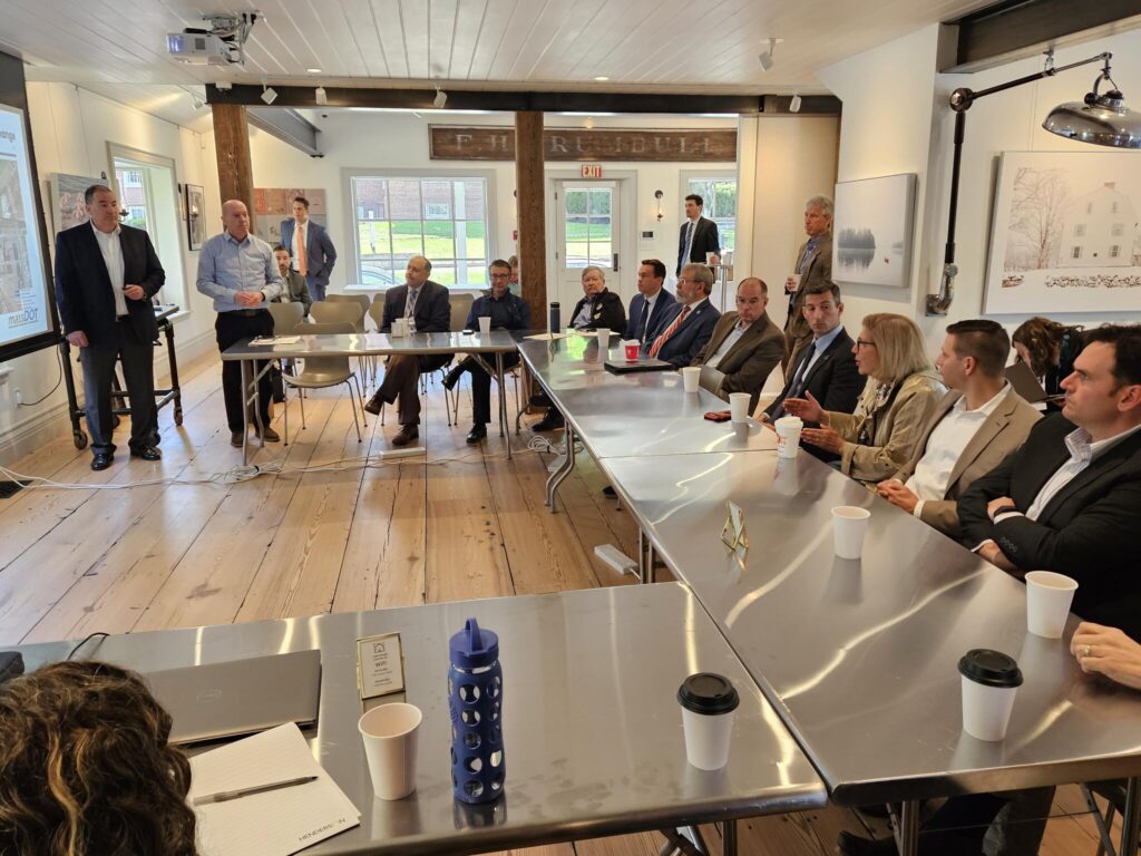 Several shiny metal tables are set in a U-shape around a screen. Every seat is full and a few people are standing in the background.