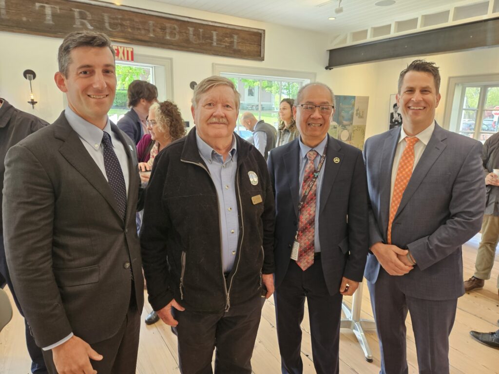 Four men stand together in a room with people in the background.