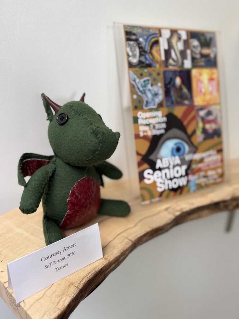 A little green handmade dinosaur? Dragon? Critter of some kind. The label idintifies it as "Self Portrait, 2026," by Courney Arnett.