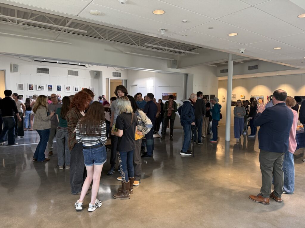 A large number of people standing in a large room. There is art on the walls and people are chatting and looking at the art.