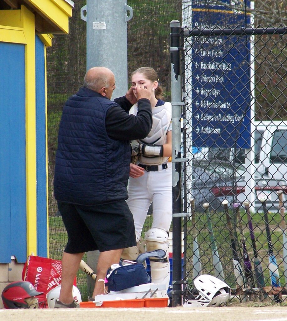 A balding man wearing a vest and shorts attends to the face of a tall young woman wearing softball catchers gear.
