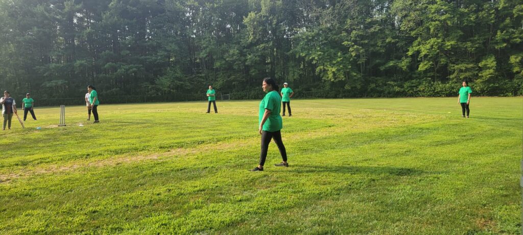Women in green t-shirts stand outdoors around a cricket wicket.