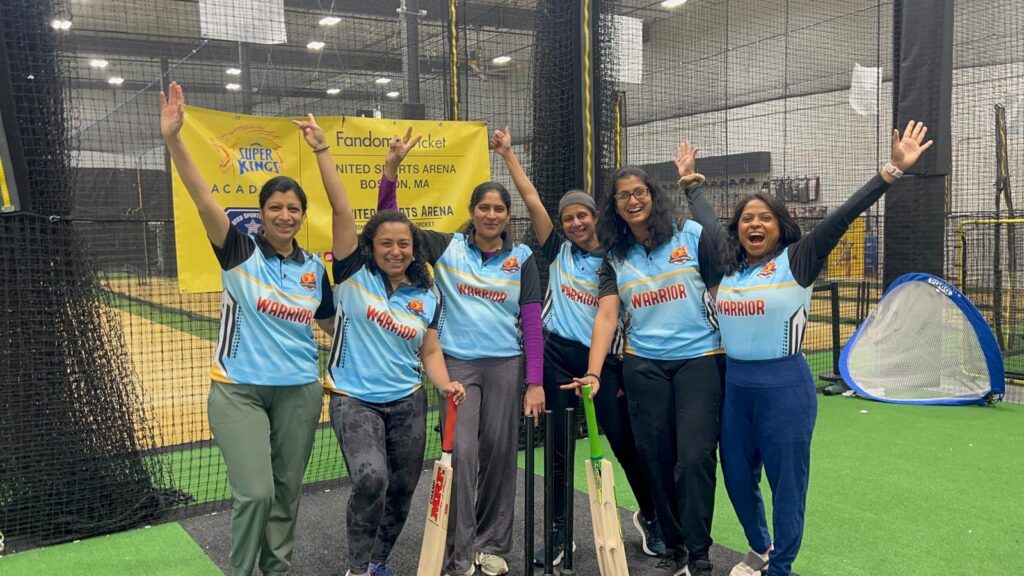 A group of south Asian women wearing blue Warrior shirts stand with their arms in the air and sport clebratory grins.