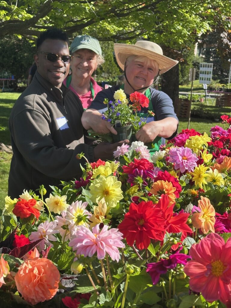 A Black man and two white women stand in front of a table full of brightly colored flowers. Two of them hold a pot that seems to have several different types of flowers in it.
