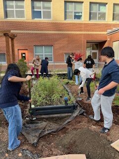 A group of teens work creating a raised bed garden in an outdoor corner of a brick building.