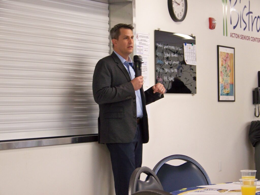 A tall man in a suit jacket holds a mic and speaks to the audience.