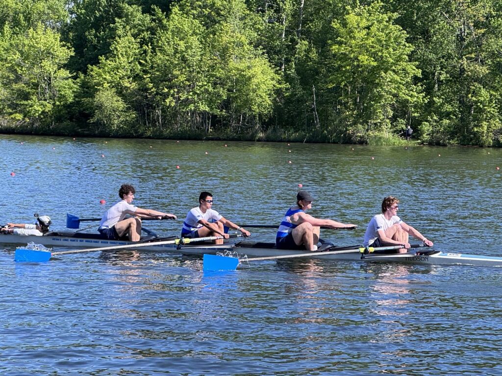 Four young men row on a sunny day. In the front, the coxswain, facing away from the rowers, sits in the well of the boat.