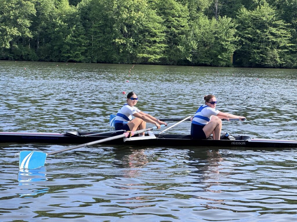 Two young women in blue and white jerseys pull the oars of a two-person boat on sparkling water.