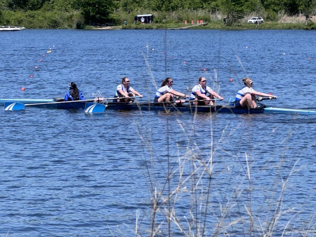 Four young women row on a sunny day. In the front, the coxswain, facing away from the rowers, sits in the well of the boat.