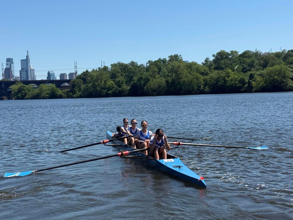 Four young women row on a sunny day. In the front, the coxswain, facing away from the rowers, sits in the well of the boat The boat is lined up with the front rower first and cox in the back (out of view)..