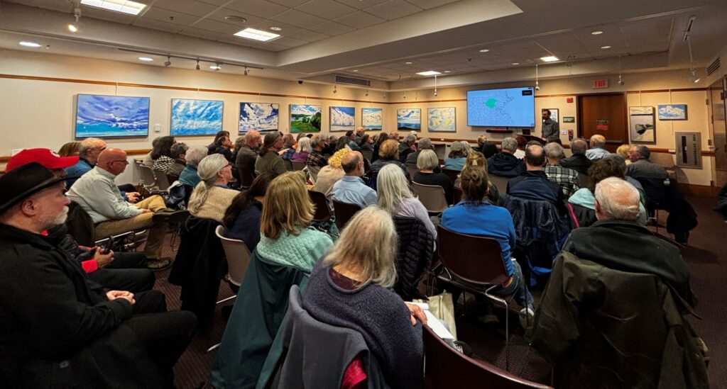 A room ful of people sitting in a room. There are blue and white painings around the room (this month's art exhibit) and a man standing at the front of the room next to a projection of a map.