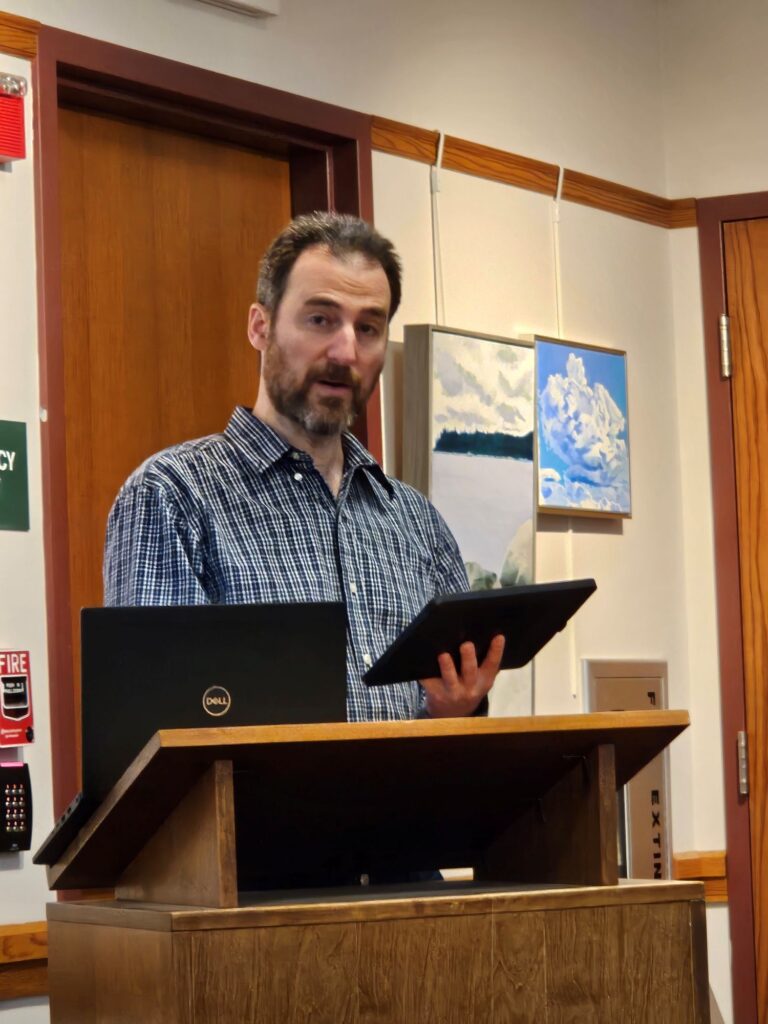 A bearded man with spiky hair and a plaid shirt holds an iPad and speacks to the audience.