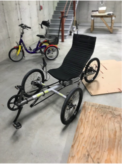 Two trikes, in the background a small upright trike for a child or small adult. In the foreground, a recumbent trike with a sling seat.