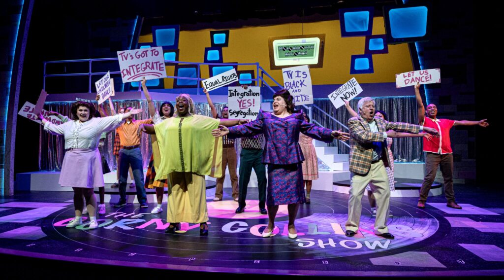 A group of people sing onstage. Some of the dancers are carrying signs that say things like:"TV's got to Integrate" and "TV is Black and White".