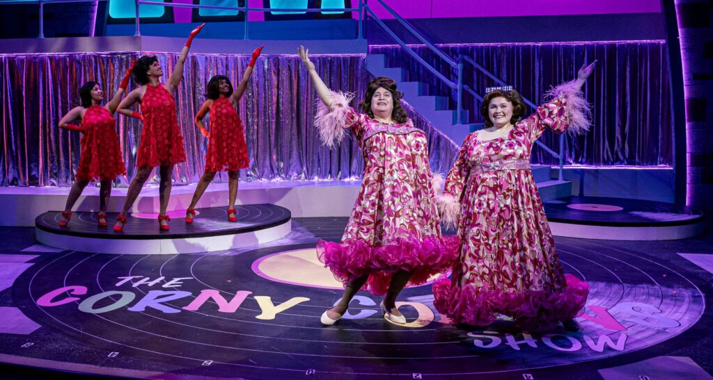 On the left, three Black women in red dresses stand with one hand on a hip and the other in the sky. On the right, two white women with big hair and fluffy pink dresses each have a hand in the air.
