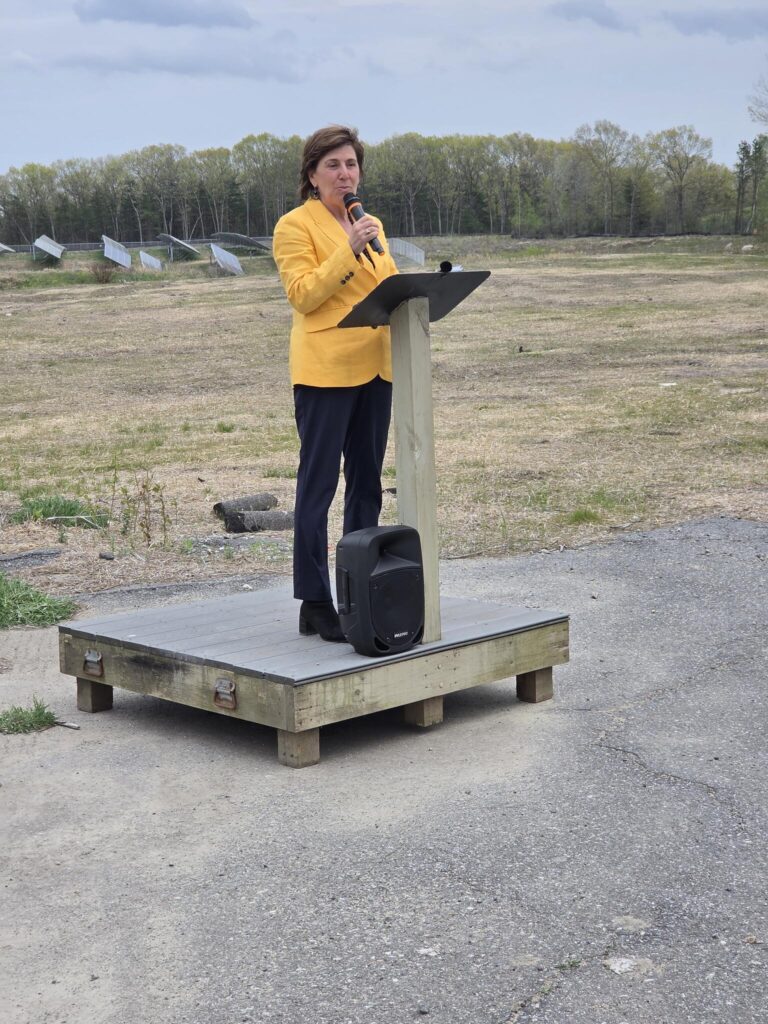 A woman in a bright yellow blazer speaks into a mic at a wooden podium. Solar panels are in the background.