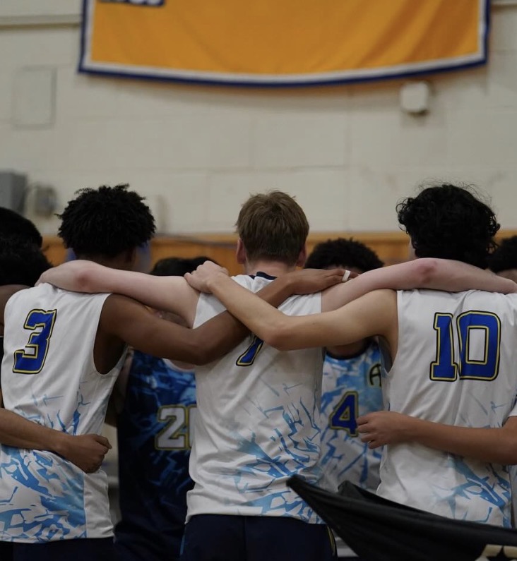 Three players from the back with their arms around each other. They are part of the larger circle and you can see lots of arms and bits of other players.
