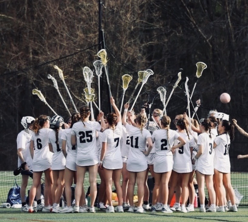 The entire team stands in a close circle with their lacrosse sticks held high.