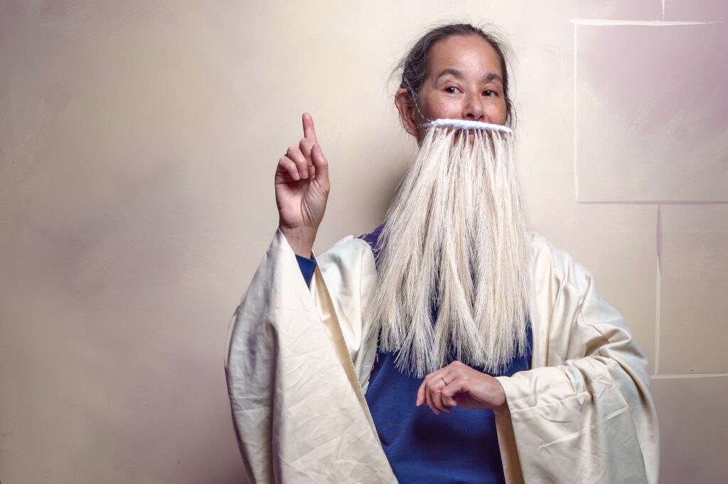 A woman wearing a long white (very fake) beard) and blue and white robes stands with one finger pointing to the sky.