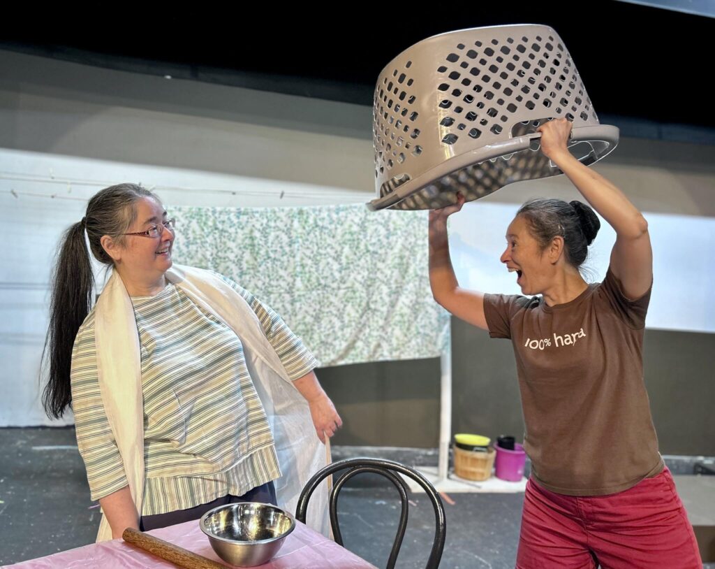 Two women stand in front of a table. The woman on the right carries a laundry basket over her head and is leaning into to the woman on the left, who is leaning away.