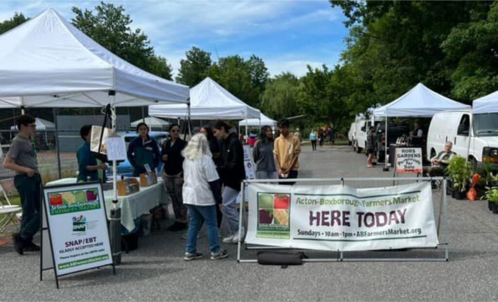 Tents in the sunshine and a big sign that says Acton-Boxborogh Farmers Market, Here Today.