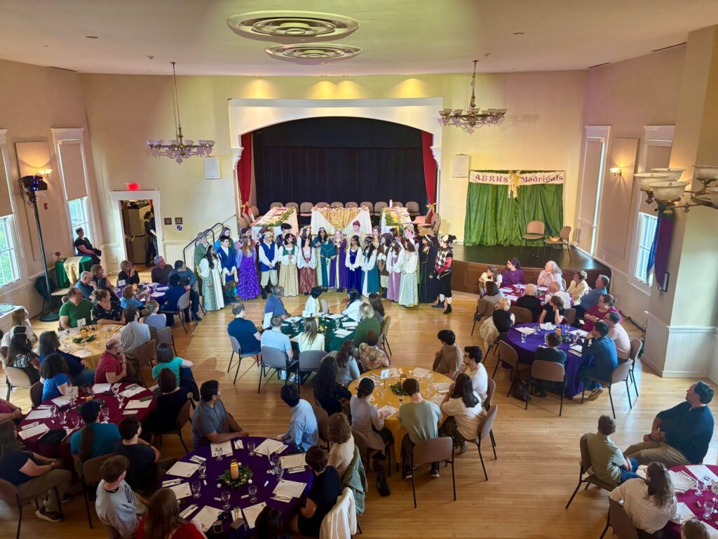 A room viewed from a balcony. At the front, a group of students in renaissance dress gathers. Around the room are tables of guests, watching and listening.