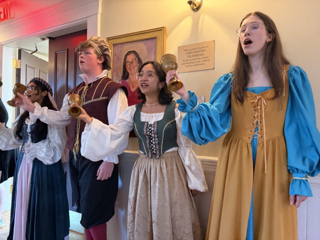 Four high-schoolers wearing (for the three girls) floor length dresses with overdresses or (for the boy), pantaloons, an embroidered vest, puffy shirt, and gold cap.