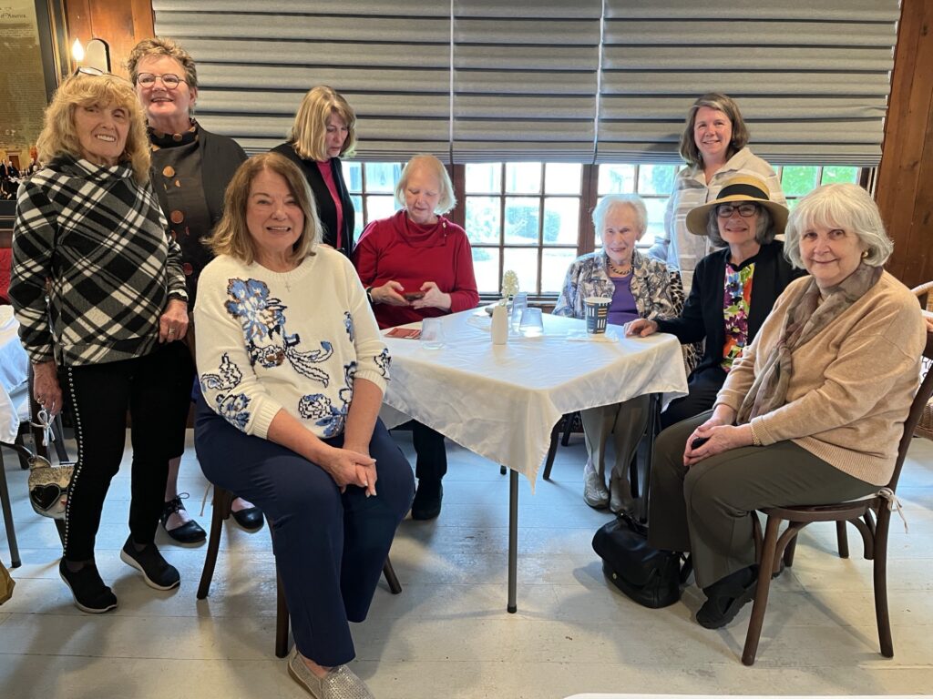A group of wemen, sit or stand around a coverd card table.
