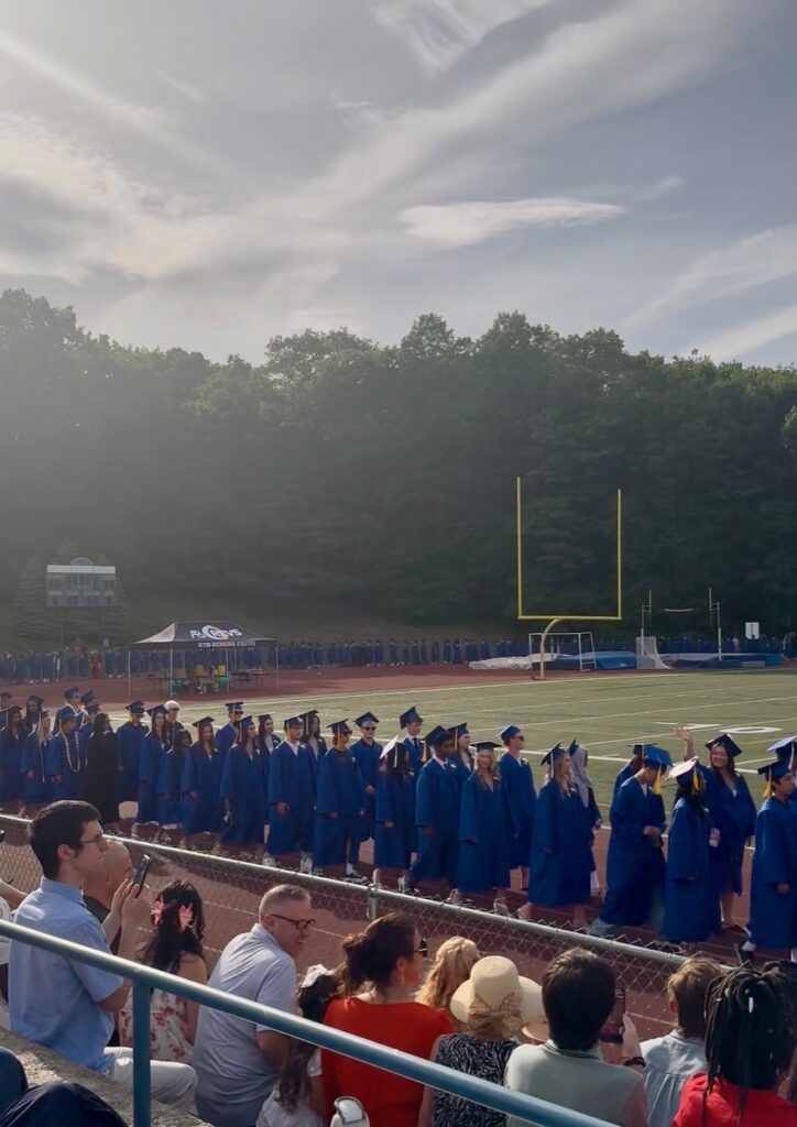 In the foreground, people sitting in the bleachers at Leary Field. In the background, graduates in blue caps and goowns are lined up in front of the bleachers and stretching back around the field.