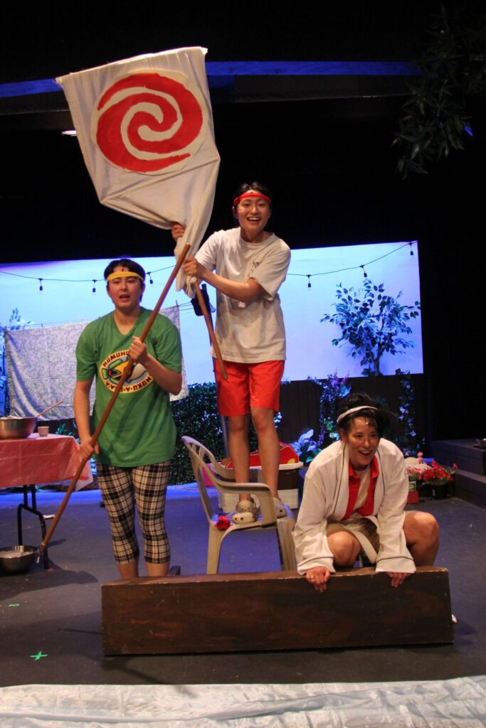 Three actors, one holding a pole, one standing on a plastic chair and holdinga colorful red and white flag, and one squatting, all behind a rectangle of brown wood about a foot high and four feet long, representing a boat. They are fully engaged and smiling.