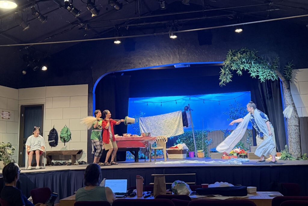 From left to right: A girl wearing animal ears sits with a book in her lap; two people next to each other walk across the stage carrying a broom and a bucket; a woman wearing a long scarf points at the broom carriers. In the background, a table, blue sky, and drying laundry